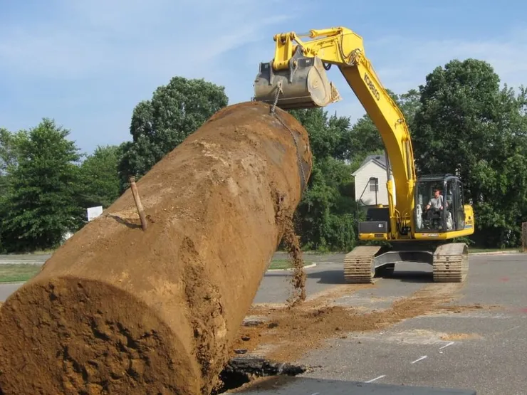 Yellow Kobelco excavator dragging a soil-covered steel underground tank from a parking-lot pit