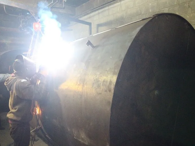 Welder fabricating a large cylindrical steel storage tank with a bright welding arc in a basement