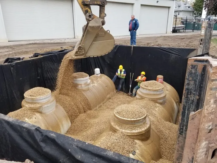 Excavator pouring pea-gravel backfill around new fiberglass tanks with crew in the pit
