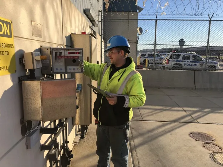 Technician in a hard hat inspecting a wall-mounted high-level alarm panel with a clipboard