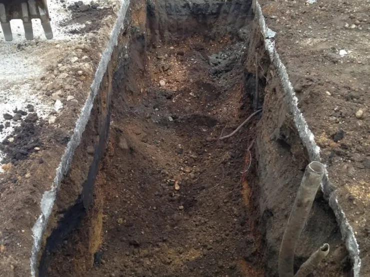 Deep rectangular excavation with exposed soil walls, a severed pipe and an excavator bucket