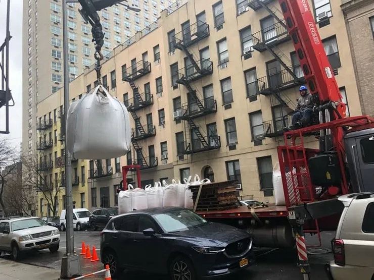 Boom truck hoisting a white bulk soil sack above a city street and flatbed trailer