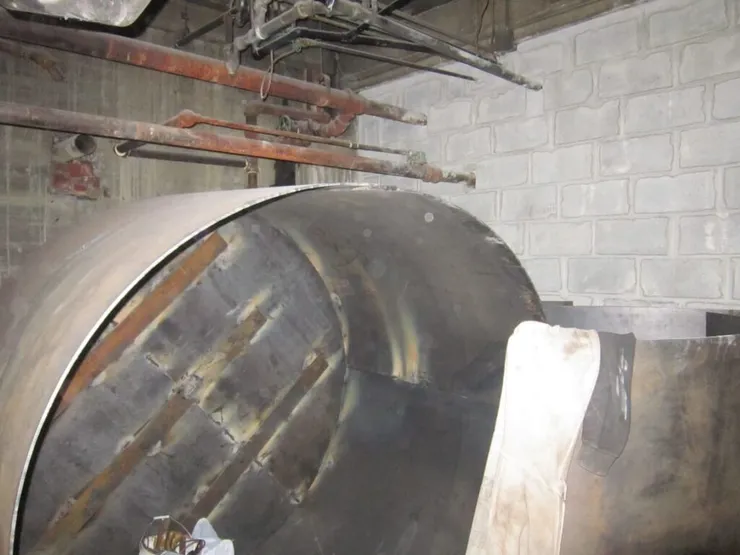 Close-up of a steel tank end being welded from plate segments below rusty basement pipes