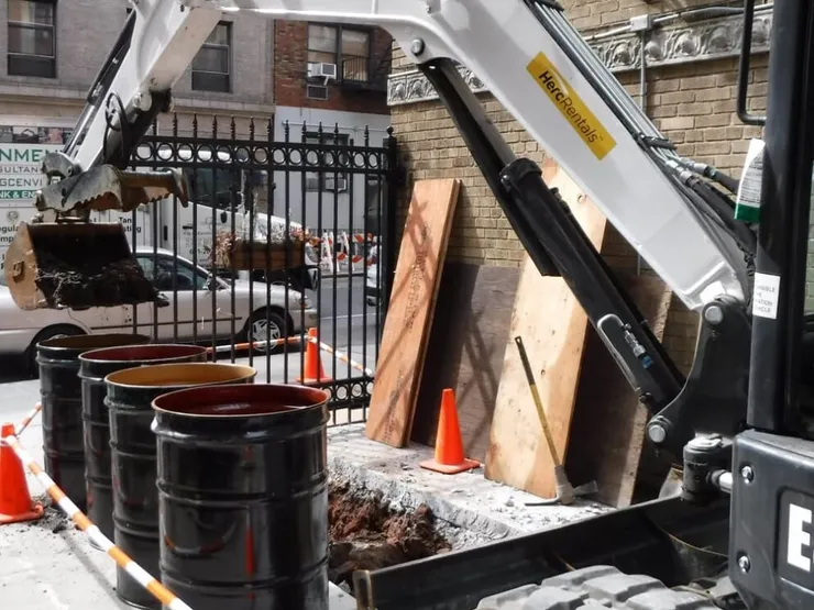 Mini-excavator dumping contaminated soil into open steel drums on a Manhattan sidewalk